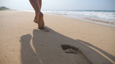 Kadın ayaklar arka planda okyanus dalgaları ile plajda altın kum üzerinde yürüyor. Genç kadının bacakları kuma basıyor. Deniz kıyısında yalınayak kız. Yaz tatili tatili. Yavaş hareket yakın çekim