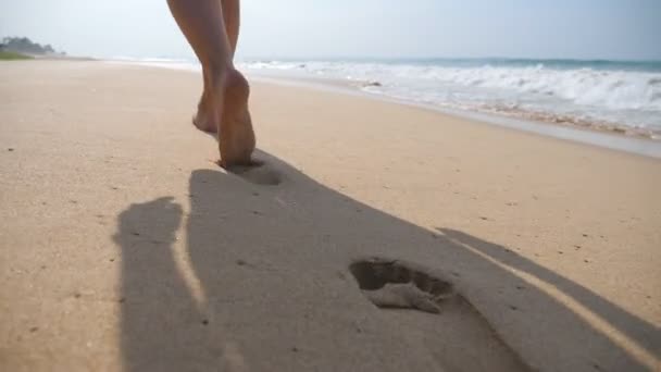 Vrouwelijke voeten lopen op gouden zand op het strand met oceaan golven op de achtergrond. Benen van jonge vrouw intensivering op zand. Barefoot meisje aan de kust. Zomervakantie. Slow Motion close-up — Stockvideo