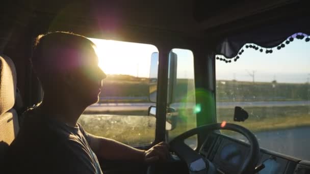Pria mengendarai truk dan hati-hati mengawasi jalan. Kaukasia pria berkuda melalui pedesaan di latar belakang matahari terbenam. Profil sopir truk di dalam taksi. Tampilan samping Lambat gerak Tutup — Stok Video