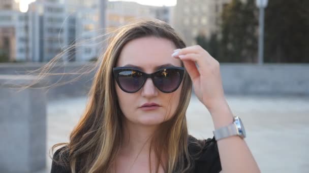 Retrato de joven empresaria en gafas de sol con paisaje urbano de fondo. Atractiva mujer de negocios mirando a la cámara y alisa el cabello. Cara de chica segura. Primer plano en cámara lenta — Vídeo de stock