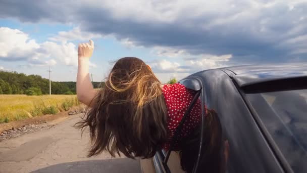 Jonge vrouw leunend uit autoraam en houden haar hand uit tijdens het rijden door landweg. Meisje kijkt uit open raam bewegende auto en rechttrekken haar lang bruin haar. Close-up achteraanzicht — Stockvideo