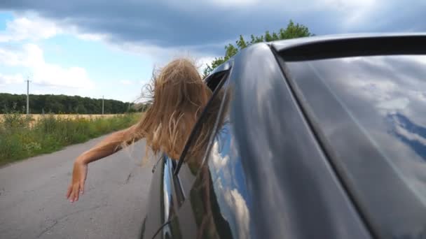 Carefree little girl leaning out of car window and waving her hand in wind while riding through country road. Small child putting her arm out of open window moving auto to feel the breeze. Slow motion — Stock Video