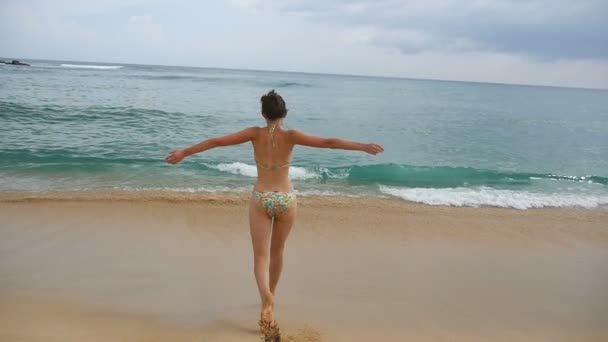 Happy woman in bikini runs towards ocean for refreshing or swimming. Young girl jogs on sand beach into the sea and enjoys freedom during vacation. Concept of summer travel. Rear view Slow motion — Stock Video