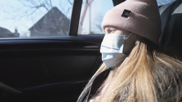 Маленька дівчинка з маскою для обличчя подорожує в машині і дивиться крізь вікно. Сумна дівчинка носить захисну маску від вірусу під час їзди. Концепція безпеки життя від пандемії коронавірусу — стокове відео