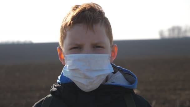 Πορτρέτο του μικρού αγοριού με ιατρική μάσκα προσώπου στέκεται σε εξωτερικούς χώρους. Θλιμμένο αρσενικό παιδί που φοράει προστατευτική μάσκα από τον ιό έξω. Έννοια της ζωής για την υγεία και την ασφάλεια από το coronavirus και πανδημία. Κλείσε. — Αρχείο Βίντεο