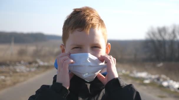 Портрет маленького мальчика в медицинской маске стоит на улице. Счастливый мальчик снимает защитную маску от вируса и улыбается. Концепция здоровья и безопасности жизни от коронавируса и пандемии — стоковое видео