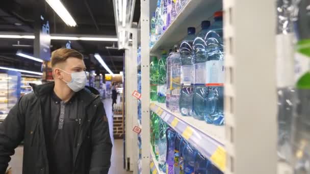 KHARKIV, UCRAINA - 18 marzo 2020: Giovane con maschera medica che sceglie e acquista una bottiglia d'acqua al supermercato. Il tipo offre da bere in negozio. Shopping in drogheria durante la pandemia di coronavirus — Video Stock