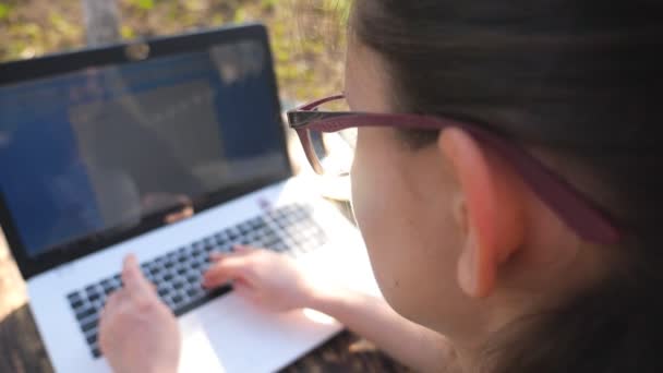 Unrecognizable businesswoman in sunglasses typing some text on keyboard of laptop. Girl using notebook outdoors. Creative female freelancer working on new project at nature. Slow motion Top view — Stock Video