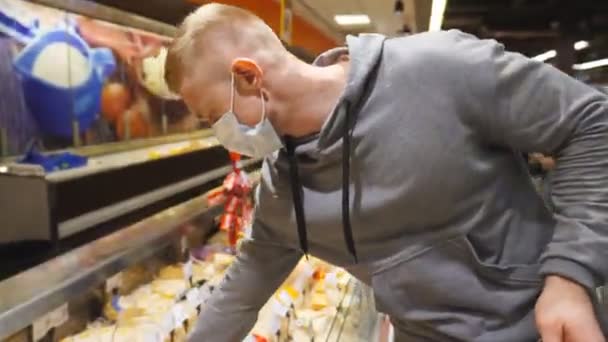 Joven con mascarilla médica está cerca de la nevera en la tienda y tomando el producto de ella. Chico seleccione el queso en la sección refrigerada en la tienda. Compras en el supermercado durante la pandemia de coronavirus — Vídeo de stock