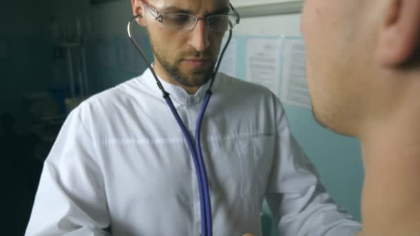 En läkarundersökning på hennes kontor på sjukhuset. Doktorn undersöker en ung manlig patient med stetoskop. Medicinsk arbetare lyssnar hjärtslag av människan. Närbild Bakifrån — Stockvideo