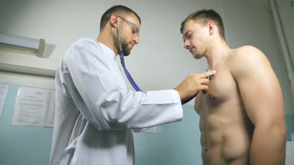 Медичний працівник слухає серцебиття людини. Медик перевіряє груди хлопця в її кабінеті в лікарні. Доктор вивчає молодого пацієнта з стетоскопом. крупним планом — стокове відео
