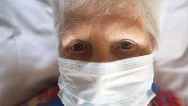 Portré egy öregasszonyról, aki védőmaszkot visel a vírustól. Beteg idős hölgy orvosi maszkkal az ágyában fekszik a kórházban. Az egészségi és biztonsági élet koncepciója a coronavirus COVID-19 világjárványtól — Stock videók