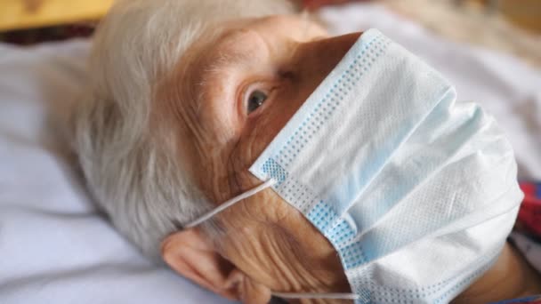 Porträtt av mormor bär medicinsk skyddsmask från virus. Sjuk dam med sorgsen syn tittar in i kameran medan han ligger på sjukhus. Begreppet hälsa och säkerhet liv från coronavirus. Närbild — Stockvideo