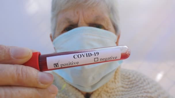 Gammel dame med beskyttende maske holder hendes blodprøve med positivt resultat på coronavirus. Syg bedstemor kigger ind i kameraet og viser triste følelser. Begrebet sundhed og sikkerhed liv fra pandemi COVID-19 – Stock-video
