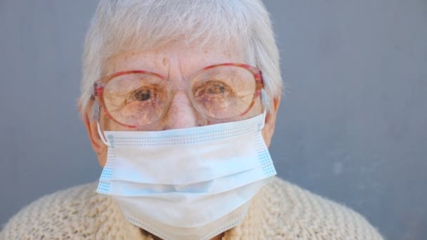 Портрет бабушки в очках носит защитную маску от вируса. Бабушка смотрит в камеру с задумчивым зрением. Концепция здоровья и безопасности жизни от пандемии. Карантин коронавируса для пожилых людей — стоковое видео