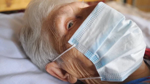 Portrait of granny wearing medical protective mask from virus. Sick lady with sad sight looking into camera while lying at bed in hospital. Concept of health and safety life from coronavirus. Close up