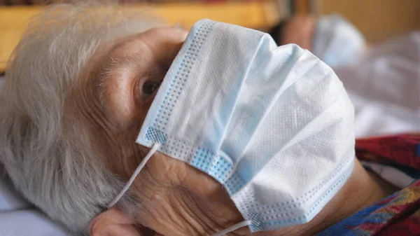奶奶头戴防毒口罩的画像 躺在医院的病床上 一位视力不好的病人看着摄像机 健康和安全生命的概念来自于头孢病毒 靠近点 — 图库照片