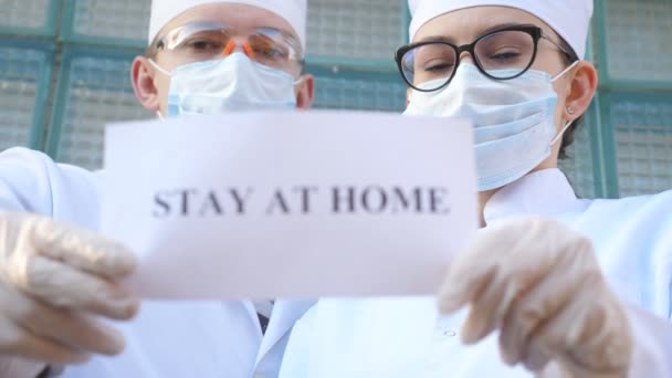 Erkek ve kadın doktorlar, Coronavirus salgını sırasında evde kalan yazıtlı bir resim tutuyorlar. Sıhhiyeciler, COVID-19 salgınından kendini izole etme ve güvenli yaşam çağrısında bulunan bir poster gösteriyor. — Stok video