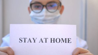 Kadın doktor, Coronavirus salgını sırasında evde kalan yazıtlı bir resim tutuyor. Sıhhiyeci kendini izole etmeye meyilli bir poster gösteriyor. COVID-19 salgınından korunma kavramı