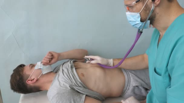 Доктор проводить обстеження пацієнта чоловічої статі зі стетоскопом, шукає симптоми COVID-19. Медик слухає легені від кашлю. Лікар перевіряє груди хлопця в лікарні. Концепція безпеки життя від пандемії коронавірусу — стокове відео