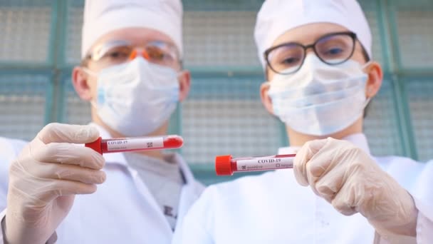 Mannelijke en vrouwelijke artsen houden reageerbuis met bloedmonster aan coronavirus. Twee dokters met beschermende handschoenen en een bril testen bloedmonsters. Concept van gezondheid en veiligheid door COVID-19 pandemie — Stockvideo