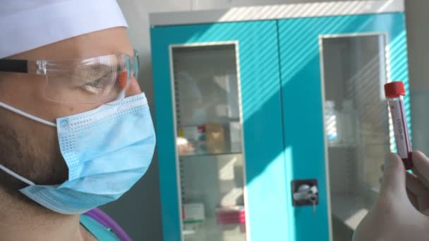 Perfil del tubo de ensayo científico con muestra de sangre a coronavirus. Trabajador de laboratorio joven en gafas protectoras examinando muestras de sangre a COVID-19. Vida útil de la pandemia del virus de la corona — Vídeo de stock