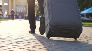 Güneşli bir günde şehir caddesinde yürüyen ve bavulu tekerlekli sandalyeyle çeken başarılı iş adamının ayakları. Kendine güvenen adam uçağına doğru gidiyor. Kentsel çevreye ayak basan ayakkabılı erkek bacakları. Yavaş çekim