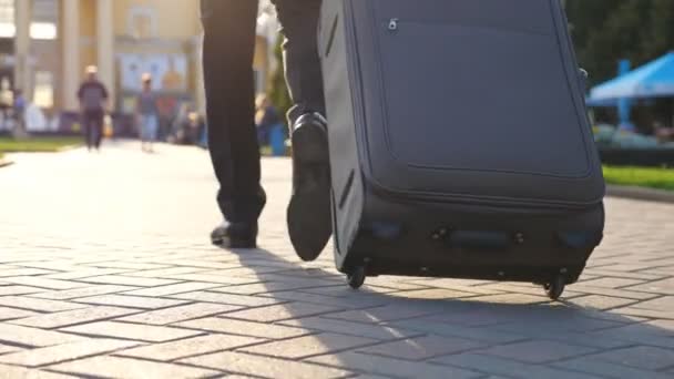 Feet of successful businessman walking through city street and pulling suitcase on wheels at sunny day. Confident man on way to his flight. Male legs in shoes stepping at urban environment. Slow mo — Stock Video