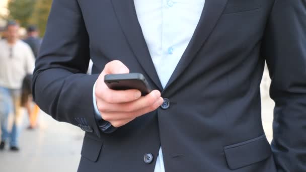 Young successful businessman in suit standing among city street and browsing smartphone. Confident man communicating or using app on his phone during break in work. Slow motion Dolly shot — Stockvideo