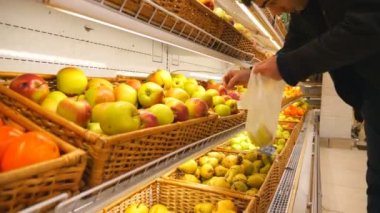 Markette taze elma seçen genç bir adam. Erkek alıcı raftan meyve alıyor ve plastik torbaya koyuyor. Süpermarkette çeşitli ürünler. Alışveriş konsepti. Yavaş çekim