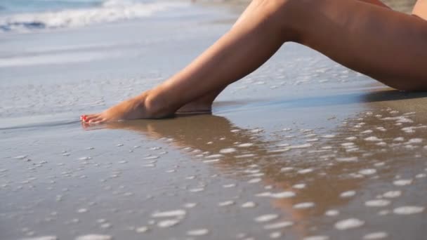 Slanke benen van een vrouw liggend op een tropisch strand met spetterende golven. Oceaangetijden spoelen over gebruinde vrouwelijke voeten. Meisje ontspannen aan de kust tijdens de zomervakantie. Zomervakantie concept. Dolly schot — Stockvideo