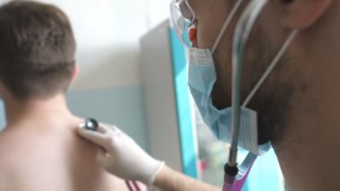 Doktor steteskoplu hastayı COVID-19 belirtileri için muayene ediyor. Sıhhiyeci insanın ciğerlerini dinler. Doktor hastanedeki adamı kontrol ediyor. Coronavirüs salgınından sağIık ve güvenlik kavramı