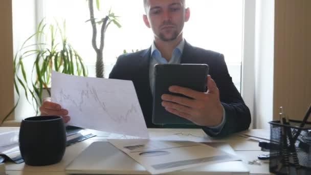 Hombre de negocios exitoso mirando diagramas con estadísticas sobre papeles mientras trabaja en dispositivos digitales. Joven emprendedor analizando la información estadística de datos en una tableta PC en el cargo. Dolly disparó — Vídeos de Stock