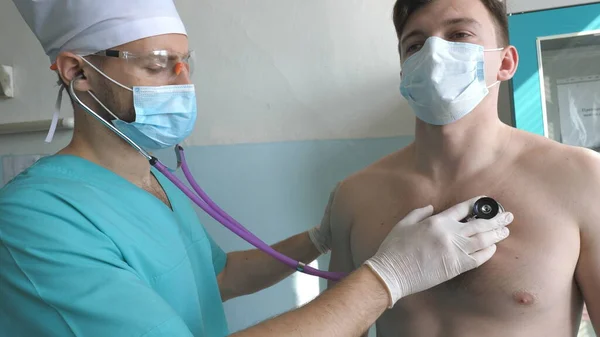 Medic with stethoscope listens lungs of patient looks for COVID-19 symptoms. Doctor with examining man with stethoscope to coronavirus pneumonia signs. Physician checking chest of guy at the hospital.