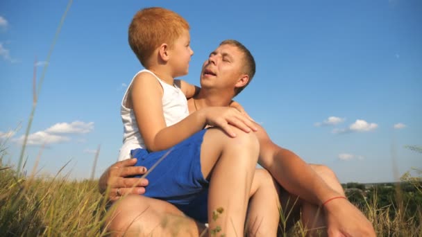 Padre sentado en la hierba verde en el césped con su chico pelirrojo y hablando entre sí. Papá y su pequeño hijo descansando en el prado. Feliz familia pasando tiempo juntos en la naturaleza. Movimiento lento — Vídeos de Stock