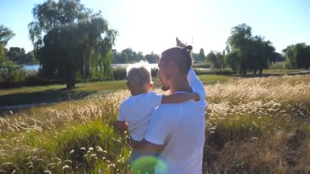 Il giovane papa 'tiene il bambino per mano sul prato. Padre e figlio che puntano al sole sul campo. Famiglia felice passare del tempo insieme. Paesaggio estivo sullo sfondo. Primo piano Retrovisore al rallentatore — Video Stock