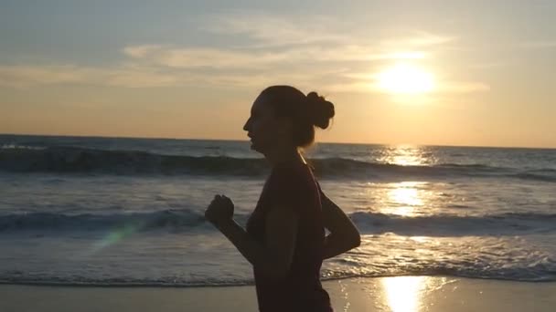 Girl jogging along ocean shore during sunrise. Silhouette of young woman running on sea beach at sunset. Female sportsman exercising outdoor. Healthy active workout lifestyle at nature. Slow motion — Stock Video
