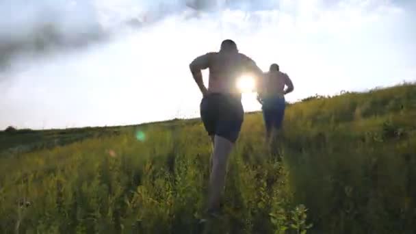 Grupo de hombres jóvenes corriendo por la colina verde sobre el cielo azul con destellos de sol en el fondo. Los atletas masculinos están corriendo en la naturaleza al atardecer. Los corredores deportivos van cuesta arriba al aire libre al amanecer. Movimiento lento — Vídeos de Stock