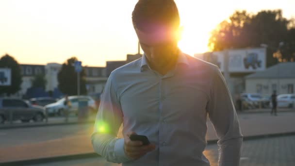 Detailní záběr mladého úspěšného obchodníka v tričku stojící v městském prostředí a používající smartphone při západu slunce. Pohledný muž píše zprávu na mobil na City Street. Sluneční světlo na pozadí — Stock video