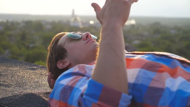 Jóképű férfi napszemüvegben a tetőn fekszik és cigizik a városkép hátterében. Fiatal srác pihentető és élvezi a szabadtéri pillanatot. Gyönyörű a kilátás. Lassú mozgás Bezárás — Stock videók