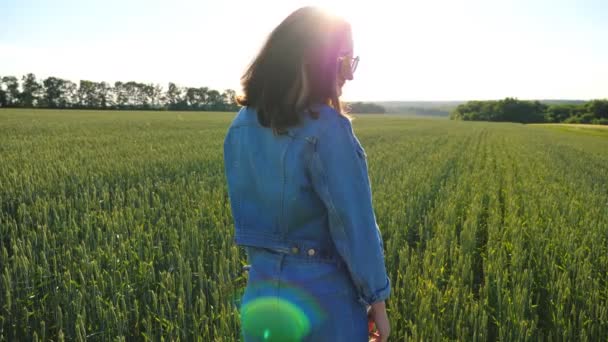 Attraktive Brünette mit Sonnenbrille, die in die Kamera blickt. Ein junges lächelndes Mädchen steht inmitten eines Feldes mit grünem Weizen und Sonnenstrahlen im Hintergrund. Schöne Landschaft. Zeitlupe — Stockvideo