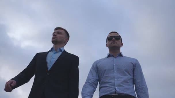 Dos jóvenes empresarios caminando por la ciudad con el cielo azul al fondo. Colaboradores guapos viajando para trabajar juntos. Colegas al aire libre. Vista de ángulo bajo Cámara lenta Primer plano — Vídeos de Stock