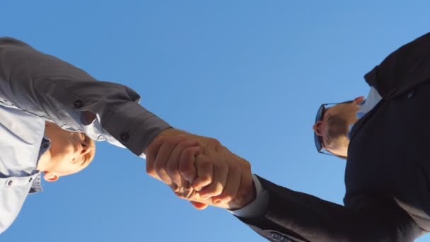 Giovani uomini d'affari si salutano e si stringono la mano su sfondo blu cielo. Due colleghi di successo se ne vanno in direzioni diverse dopo essersi incontrati all'aperto. Movimento lento Vista ad angolo basso — Video Stock