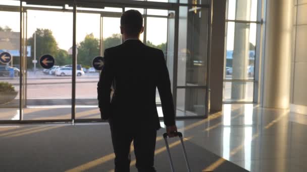 Unbekannter Mann geht mit seinem Gepäck durch gläserne automatische Tür des modernen Flughafens in Richtung Stadtstraße. Geschäftsmann, der vom Terminal zu den Parkplätzen läuft und Koffer auf Rädern zieht. Zeitlupe — Stockvideo