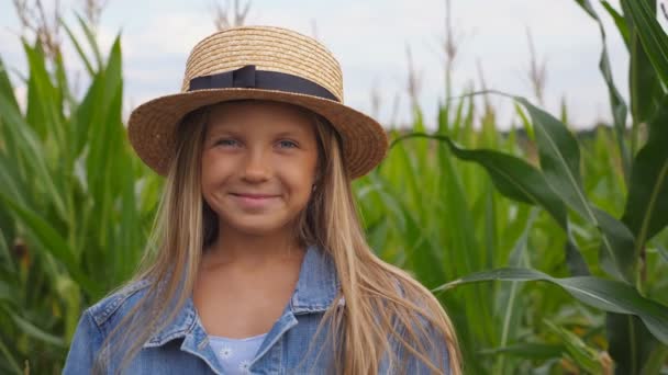 Pequeña chica divertida en sombrero de paja mirando a la cámara y se ríe cubriendo su cara con la mano en el campo de maíz. Retrato de una niña feliz jugando con su largo cabello rubio y alisando su sombrero de paja — Vídeos de Stock