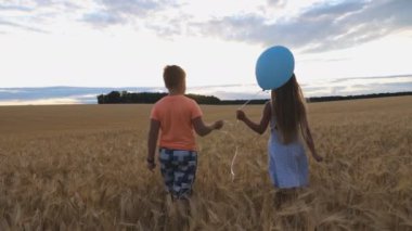 Küçük kız ve oğlan ellerinde balonla gün batımında buğday tarlasında yürüyorlar. Bulutlu bir günde arpa tarlasına giden iki küçük şirin çocuk. Çocuk sevgisi kavramı. Yavaş çekim