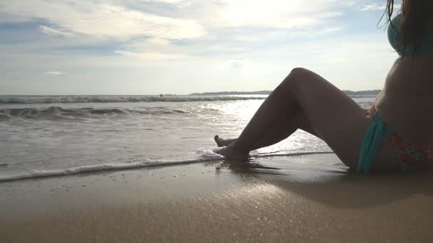 비키니를 입은 소녀는 해안에 앉아 햇빛 이 잘 드는 날에 일광욕을 한다. 아름다운 여자가해 안에서 휴식을 취하고 아름다운 경치를 감상 한다. 여름 방학이나 휴일의 개념. 돌리 샷 슬로우 모션 — 비디오