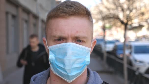 Retrato de jovem com máscara facial médica fica na rua da cidade. Um tipo a usar máscara protectora do vírus ao ar livre no meio da multidão. Conceito de saúde e segurança da pandemia de coronavírus — Vídeo de Stock