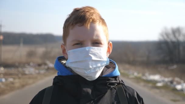 Porträt eines kleinen Jungen mit medizinischer Gesichtsmaske, der im Freien steht. Trauriges männliches Kind mit Schutzmaske vor dem Virus draußen. Konzept der Gesundheit und Sicherheit vor Coronavirus und Pandemie. Nahaufnahme — Stockvideo