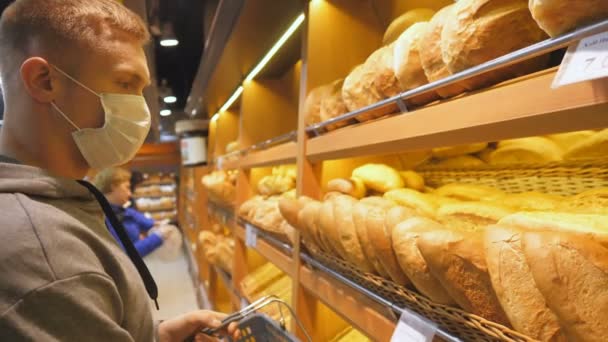 Junger Mann mit medizinischer Gesichtsmaske wählt im Supermarkt ein frisches Brot. Guy, der zur Quarantänezeit Lebensmittel in der Lebensmittelabteilung auswählt. Einkaufen im Lebensmittelgeschäft während der Coronavirus-Pandemie — Stockvideo